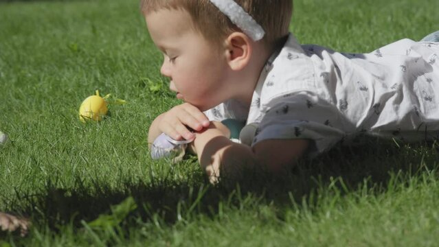 happy smiling Easter boy portrait child lying on fresh green grass outdoors in park wearing Easter buny ears grab colorful Easter eggs lying on stomach on fresh spring grass. Easter egg hunt game