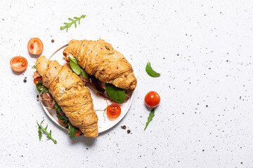Fresh aromatic full grain croissant with jamon or prosciutto, eggs, cheese, tomatoes and salad leaves on white marble background close up. Tasty and healthy breakfast idea.