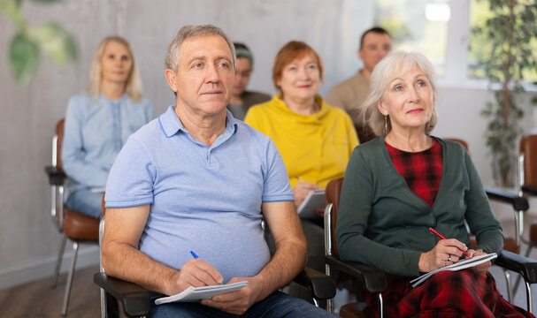 Positive interested elderly man listening attentively and taking notes on lesson during language courses for older adults