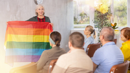 Smiling elderly female tutor participating in seniors education program, talking about international LGBT social movements to group in auditorium, showing colorful rainbow flag..