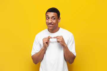 young bashful african american guy in white t-shirt is shy on yellow isolated background,...