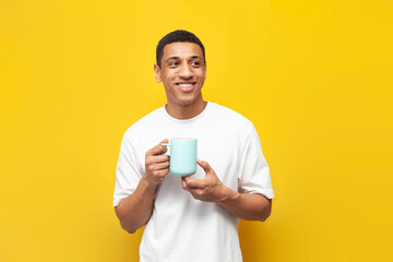 young african american guy in white t-shirt holds cup of coffee on yellow isolated background, man...