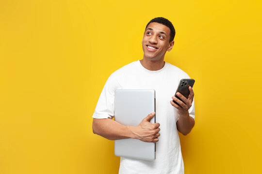 young african american guy in white t-shirt holding laptop and using smartphone and dreaming on yellow background