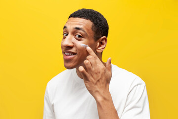 american guy in white t-shirt applies cream on his face on yellow isolated background, man uses...