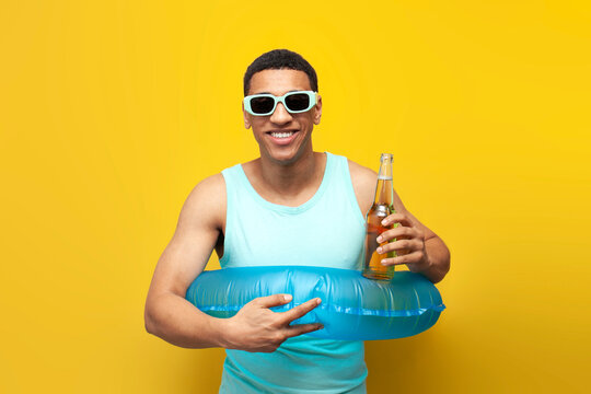 cheerful african american guy in sunglasses with inflatable swimming circle holds bottle of beer on yellow background - Powered by Adobe