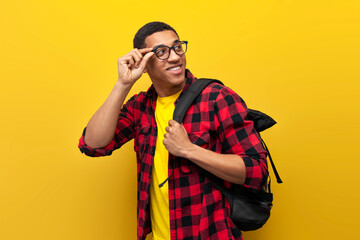 young guy african american student in glasses with backpack looks back on yellow isolated background