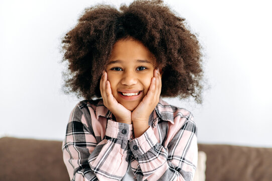 Photo of a cute beautiful African American preschool girl, with curly hair, in a basic clothes, sits on the sofa, looks to the camera, smiles friendly. Happy childhood