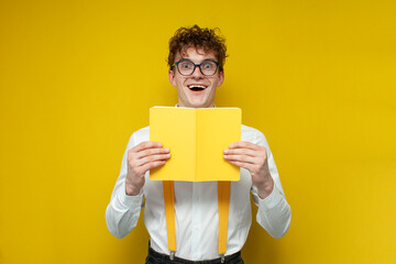 funny nerd guy in glasses holds book and wonders on yellow isolated background, student in festive...