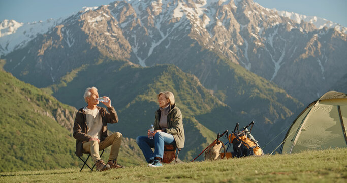 Old caucasian couple taking a rest on top of mountain, sitting and rehydrating while enjoying the view, talking. Mature people spending time together after retirement travelling - tourism, pension