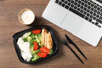 Container with tasty food, laptop, cutlery and cup of coffee on wooden table, flat lay. Business lunch