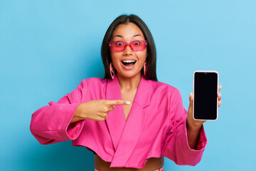 Naklejka premium Young woman in pink sunglasses and jacket points finger at her phone in surprise, girl standing isolated on blue background, big discounts in favorite market, copy space, high quality photo