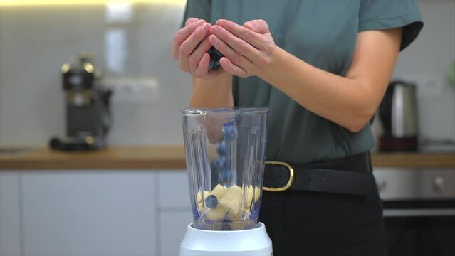 Woman putting fresh blueberries in blender with banana pisces for smoothie