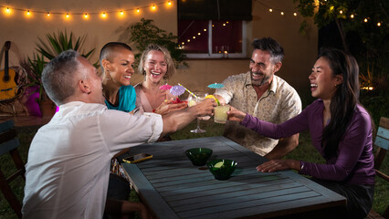 Diverse group of friends laughing and having fun during night garden party drinking cocktails.