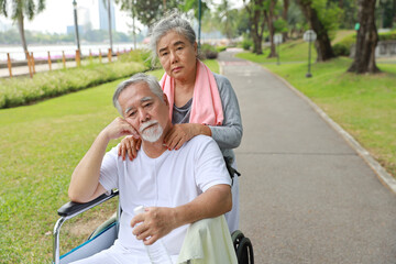 Asian senior woman or caregiver helping and consoling senior man walk with wheelchair at park outdoor. Elderly wife taking good help care and support of elder husband patient outside the house.