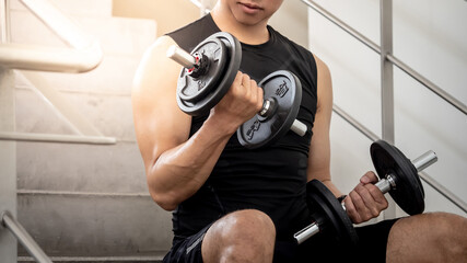 Sport man with well trained body in black sportswear sitting on concrete stair steps wearing sport gloves and lifting dumbbells. Weight training and bodybuilding workout concept.