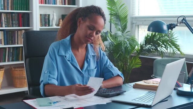 Young concentrated ethnic African American woman auditor reconciles financial accounts by checking checks and using calculator to find extra expenses to optimize budget sits at office desk with laptop