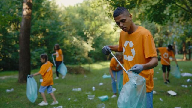 Group of Diverse Volunteers In T-Shirts with Recycling Symbol Cleaning Up the Public Park. Multiethnic Man Activist Using Trash Picker While Collecting Garbage in Bin Bag. Safe Ecology Concept