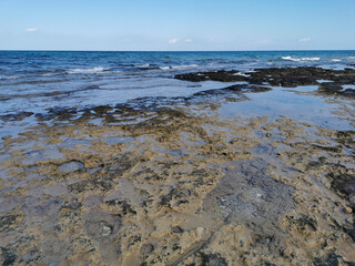 The rocky coast of the Mediterranean Sea from long-hardened lava, waves, clear water against a blue sky with clouds.
