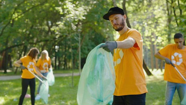 Caucasian Bearded Man Eco Volunteer Using Trash Picker While Collecting Plastic in Bin Bag. Group of Eco Activists in T-Shirts with Recycling Symbol Cleaning Park. Safe Ecology Concept