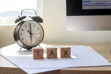 The word tax on wooden cubes with an alarm and tax forms on desk. Tax payment reminder or annual taxation concept.