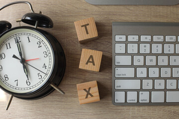 The word tax on wooden cubes with an alarm and keyboard clock on wooden desk. Tax payment reminder or annual taxation concept.
