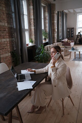 Happy businesswoman talking on mobile phone while analysing documents in her laptop.