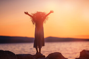Child stands on seashore at sunset with his hands raised to sky. Golden orange sunlight. Happy holidays by sea concept.