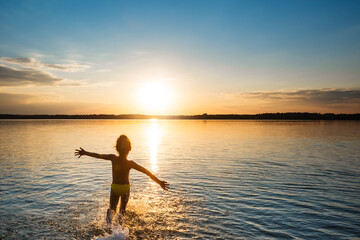 Happy child runs splashing water on beach towards the sun. Back view, wide angle shooting.