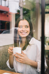 Vertical portrait of lovely woman in white shirt holding a cup of coffee. Young black haired girl with red lipstick posing