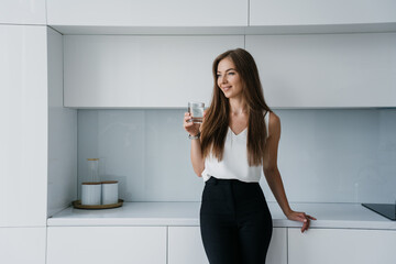 Attractive fit businesswoman in elegant clothes standing at kitchen holds glass of water looks aside smiles. Cheerful Italian female satisfied by healthy lifestyle. Successful entrepreneur dreaming.