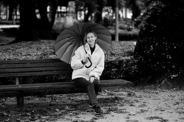 A woman sitting with an umbrella on a park bench, looking at the camera. Black and white photo.