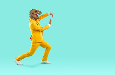 Fototapeta na wymiar Cheerful and eccentric man in rubber dinosaur mask shows funny dance moves near copy space. Man in yellow bright suit and absurd animal mask on his head isolated on turquoise background. Full length.