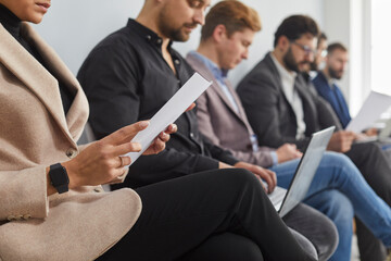 Serious young business people stylishly dressed. Group of candidate are sitting on chairs in a queue, waiting for an interview, holding questionnaires, laptops and mobile phone. Employment concept.