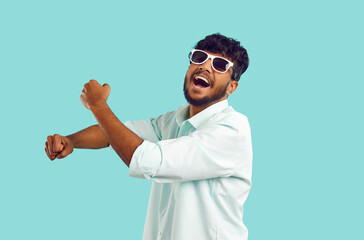 Cheerful young Indian man laughing, singing, having fun and dancing against light blue background....