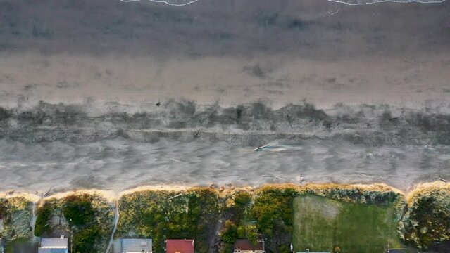 Slow top-down aerial pan from beachfront houses and green slope, across highly textured beach, to rolling waves.  Rockaway Beach, Oregon