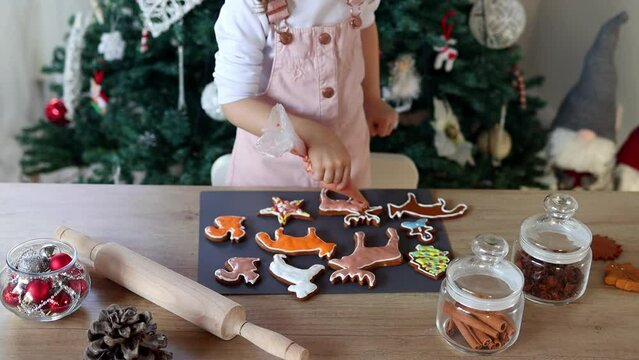 Happy little girl coloring Christmas gingerbread cookies at home. Christmas decoration in the kitchen. Fir tree with fairy lights. The concept of the New Year and Christmas. Holiday atmosphere.