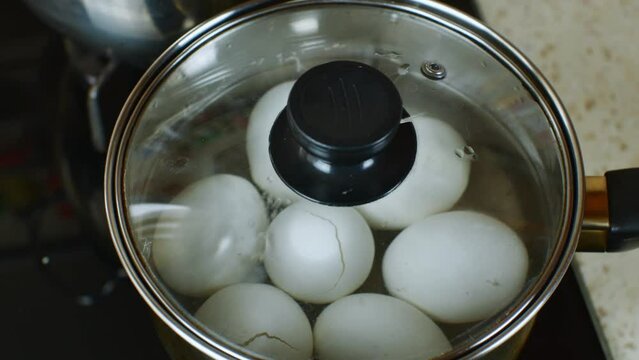 White chicken eggs are boiled in a stainless steel iron saucepan with transparent glass lid. The female hand closes a pan with boiled white chicken eggs with transparent glass roof. Close-up top view