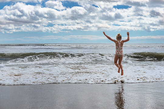 Rear view of playful girl jumping in waves at Virginia Beach against cloudy sky