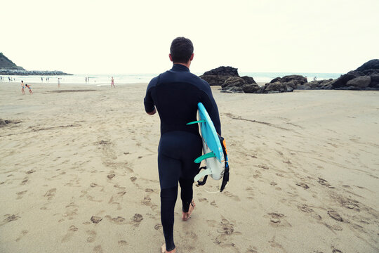 Rear view of man in wetsuit carrying surfboard while walking on beach