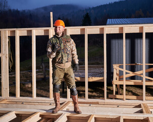 Male worker building wooden frame house. Man standing on construction site in orange safety helmet, gesturing rock and roll symbol, showing obscene horns gesture.