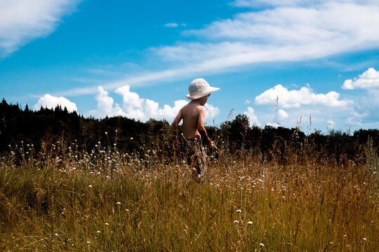 Rear view of shirtless boy wearing hat while standing on field against cloudy sky