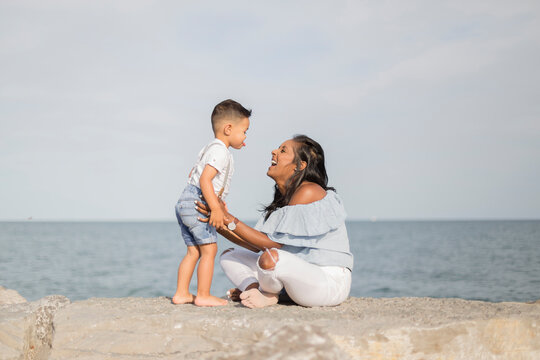 Side view of happy son and mother playing while sitting on retaining wall against sea