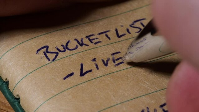 Person writing self help bucket list goal setting to live aim on brown lined paper journal, close up
