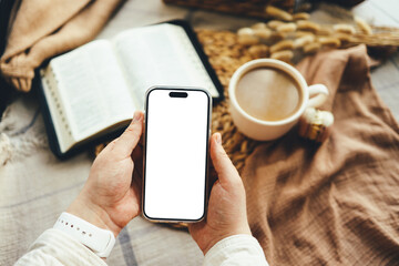 Hands holding a phone with an isolated screen, seen from above, with an open Bible, a coffee cup, and macarons in a home interior