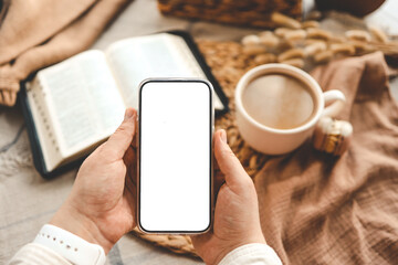 Hands holding a phone with an isolated screen, seen from above, with an open Bible, a coffee cup, and macarons in a home interior