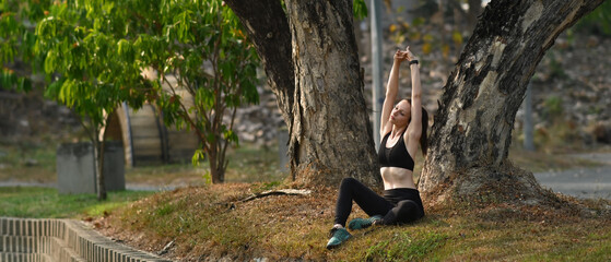 Horizontal full length view of athletic caucasian woman stretching arms, resting after workout on grass in summer park