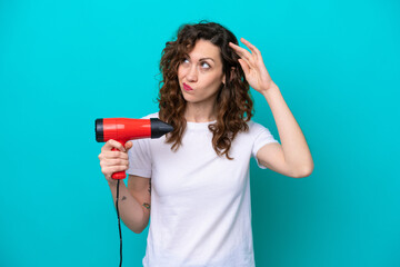 Young caucasian woman holding a hairdryer isolated on blue background having doubts and with confuse face expression