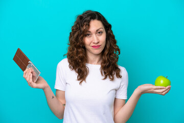 Young caucasian woman isolated on blue background having doubts while taking a chocolate tablet in...