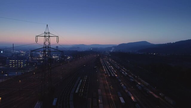 Hochspannungsleitung mit Strommast. Morgengrauen im Industriegebiet mit rauchende Schornsteinen von Fabriken, G&uuml;terverkehr und viele Bahngeleise. 