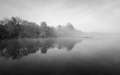 Jetty at the lake in the fog. Misty landscape in the morning. Idyllic nature by the water. Rest and relaxation.
